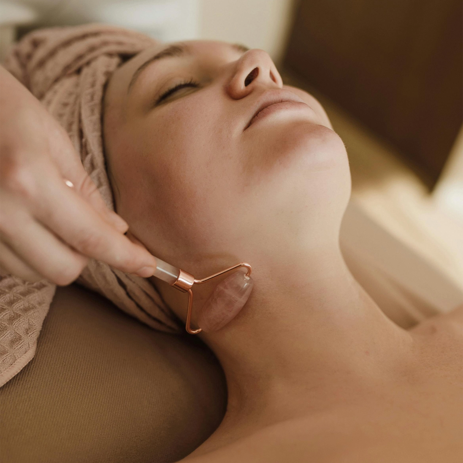 Woman lying down with a sand coloured towel around her head, receiving a facial treatment with a face roller